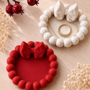 May include: Two circular trinket dishes, one solid red and one speckled white, each featuring a decorative bow and a border of small spheres. The white dish contains a gold-toned ring and a thin gold chain, while the red dish is empty.