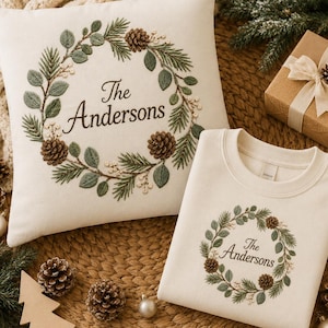 May include: A square off-white throw pillow and a matching off-white crewneck sweatshirt, both featuring an embroidered wreath design of green pine needles, leaves, and brown pinecones with the text The Andersons centered in the middle.