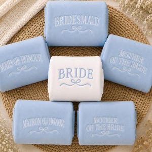 May include: A collection of six folded rectangular blankets featuring embroidered text and a decorative flourish. One white blanket is labelled BRIDE, while five light blue blankets are labelled BRIDESMAID, MAID OF HONOUR, and MOTHER OF THE BRIDE.