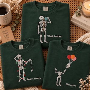 May include: Three dark green crewneck sweatshirts featuring embroidered skeleton graphics. One shows a skeleton drinking coffee with the text "That tracks.", another shows a skeleton tangled in Christmas lights with "Festive enough.", and the third shows a skeleton losing its head to balloons with "Not again."