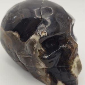 Puede incluir: Una escultura de calavera tallada con una superficie pulida. La calavera es principalmente negra con áreas de veteado marrón y crema. Una grieta atraviesa la parte superior de la calavera. La escultura es un objeto decorativo.
