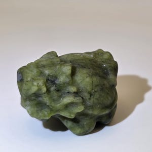 May include: A small, hand-carved figurine of a frog made from a mottled olive-green stone. The sculpture features a textured surface with visible natural veining, dark inclusions, and defined facial features including eyes and a wide mouth.