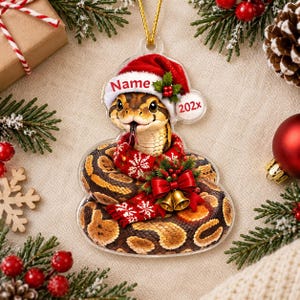 May include: A flat, clear acrylic Christmas ornament shaped like a coiled snake wearing a red and white Santa hat with the text Name and 202x. The snake is wrapped in a red and white snowflake patterned scarf and adorned with a red bow and gold bells.