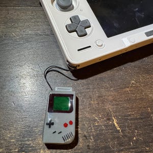 May include: A gray miniature Game Boy pendant with a green screen and red buttons is connected to a white handheld gaming console. The pendant is a detailed replica of the classic handheld console.