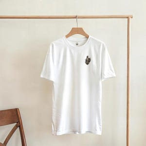 Peut inclure: Un t-shirt blanc avec un petit écusson détaillé sur la poitrine. L'écusson représente une figure dans des tons marron et gris. Le t-shirt est suspendu à un portant en bois. Le t-shirt est fait d'une matière douce et confortable.