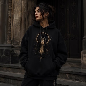 Puede incluir: Sudadera con capucha negra con un diseño detallado de una mujer con cabello largo y oscuro, una corona y un diseño celestial. La obra de arte está centrada en la parte delantera de la sudadera, con un halo dorado y detalles de estrellas.