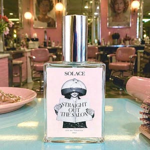 May include: A rectangular glass perfume bottle with a silver cap. The label features the text SOLACE, STRAIGHT OUT THE SALON, and EAU DE TOILETTE 30ml, with a black and white illustration of a person wearing a hooded hair dryer and reading a magazine.