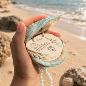 Può includere: Un taccuino decorativo a forma di conchiglia, aperto per rivelare una pila di pagine rotonde. Il guscio è turchese e bianco, con una collana di perle visibile. Le pagine del taccuino hanno illustrazioni e testo.