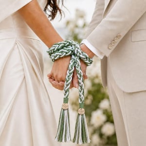 May include: A braided handfasting cord made of sage green and white yarn, wrapped around two joined hands. The cord features two hanging tassels, each finished with a small circular wooden charm engraved with text and a heart symbol.