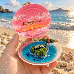May include: A hinged seashell ring box featuring a hand-painted pink sunset scene with the text Marry me on the lid. The interior contains a miniature island diorama with blue resin water, two small black whales, and a silver leaf-shaped ring.