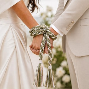 May include: A handfasting cord featuring a braided design in olive green, white, and natural tan fibers. The cord is wrapped around two joined wrists, with two long, loose ends hanging down, each finished with a thick, matching decorative tassel.