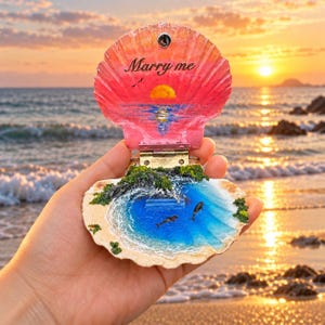 May include: A hinged seashell ornament featuring a painted sunset scene with the text Marry me on the top half and a three-dimensional coastal landscape with blue water, green foliage, and two small whale figures on the bottom half.
