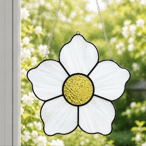 May include: A stained glass suncatcher shaped like a five-petaled flower with white textured glass petals and a bright yellow, pebbled glass centre. The piece is framed in black metal and hangs from a silver-toned metal chain.