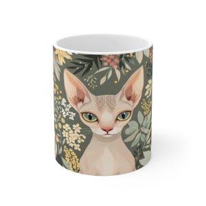 May include: A ceramic mug featuring a light-skinned, hairless Sphynx cat with large, pointed ears and green eyes. The cat is centered against a dark olive green background decorated with a pattern of small yellow, white, and pink flowers and leaves.