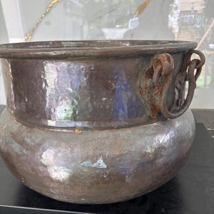 May include: A hammered copper pot with a wide rim and rounded body. The pot features two ornate, rusty metal handles on either side. The copper has a dark, aged patina, with visible hammer marks across the surface. The pot is likely an antique or vintage piece.