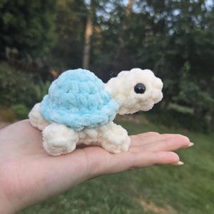 Op de afbeelding: Een kleine, handgemaakte gehaakte schildpad knuffel met een lichaam van zacht wit garen en een schild van lichtblauw garen. De schildpad heeft een ronde kop met één zichtbaar zwart plastic oog, vier korte pootjes en een pluizige textuur.