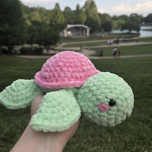 Op de afbeelding: Een pluche schildpadspeeltje gemaakt van zacht, getextureerd garen. De schildpad heeft een afgerond, felroze schild en een licht mintgroene kop, lichaam en flippers. Het heeft kleine, zwarte kraaloogjes en twee piepkleine, roze vlekjes op de wangen.
