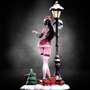 以下が含まれることがあります： 赤と白のサンタ衣装を着た女性のクリスマスをテーマにしたフィギュア。雪に覆われた街灯に寄りかかっている。プレゼントとクリスマスツリーが土台に。街灯にはリースと雪が積もっている。