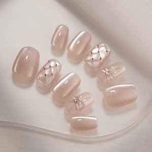 May include: A collection of light pink artificial nails. Some nails have a quilted design with small, gold-colored embellishments. Other nails feature a bow design with silver accents. The nails are arranged on a white surface.