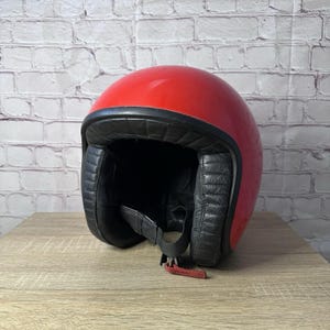 May include: A bright red open-face motorcycle helmet featuring a smooth, glossy outer shell and a black rubber trim along the edge. The interior is lined with black quilted padding and includes a black chin strap with a small red fabric pull tab.