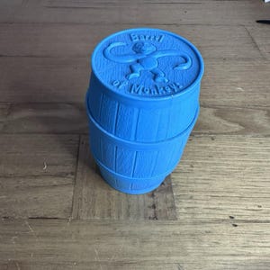May include: A bright blue plastic Barrel of Monkeys game container with a cylindrical shape and vertical wood-grain texture. The lid features a raised relief of a monkey figure and the embossed text Barrel of Monkeys in a circular arrangement.