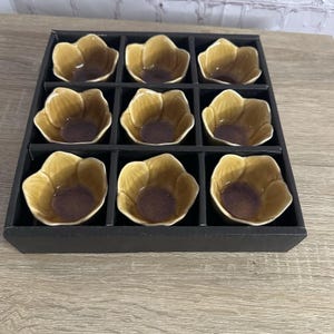 Puede incluir: Un conjunto de nueve cuencos de cerámica dispuestos en una bandeja negra con cuadrícula cuadrada. Cada cuenco tiene forma de flor con pétalos curvos, acabado en esmalte amarillo mostaza brillante y un tono púrpura intenso en la base.
