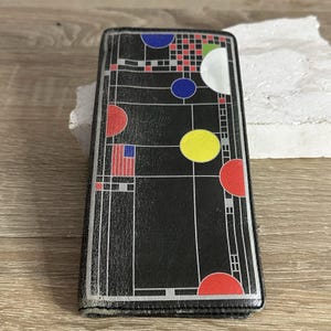 May include: A rectangular black wallet featuring a geometric grid pattern with thin white lines. The design includes solid circles in blue, yellow, and red, along with small red and blue square accents and a white semi-circle shape on the surface.