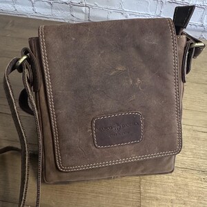 May include: A brown messenger bag with a flap closure, featuring a rectangular patch embossed with the text MARCO DANIELA COLLECTION. The bag has visible light-coloured stitching along the edges and an adjustable shoulder strap with metal hardware.