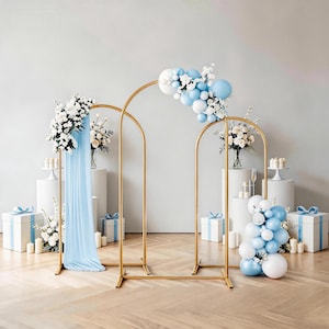 May include: Three gold metal arch frames arranged in a tiered display. The arches are decorated with clusters of light blue and white balloons and white floral arrangements. A light blue fabric drape hangs from the left arch, and white gift boxes.
