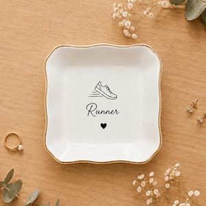 May include: A square white ceramic trinket dish with scalloped edges and a gold-painted rim. The centre features a black line drawing of a running shoe, the word "Runner" in a cursive script, and a small black heart icon below the text.