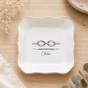 May include: A square white ceramic trinket dish with scalloped edges and a gold-painted rim. The center features a black line drawing of swimming goggles, a blue beaded line, and the name Chloe written in black cursive script.