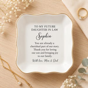 Peut inclure: Un vide-poche carré en céramique blanche avec des bords festonnés et un rebord doré. Le centre présente un texte imprimé en noir : TO MY FUTURE DAUGHTER IN LAW Sophia You are already a cherished part of our story. Thank you for loving our son and bringing joy to our family. With love, Mom & Dad.