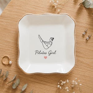 May include: A square white ceramic trinket dish with scalloped edges and a gold-toned rim. The centre features a black line drawing of a person in a Pilates pose, the text "Pilates Girl" in black script, and a small pink heart icon below.