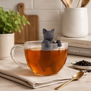 Op de afbeelding: Een donkergrijze siliconen thee-ei in de vorm van een kat, met de voorpoten rustend op de rand van een doorzichtige glazen theekop gevuld met amberkleurige thee. Het onderste deel van de katvormige infuser is ondergedompeld in de vloeistof.