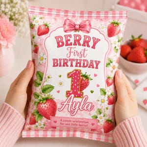 May include: A rectangular party favor bag with a pink gingham border, featuring a strawberry theme with illustrations of strawberries and white flowers. The text reads BERRY First BIRTHDAY, the number 1, the name Ayla, and A sweet celebration for our little berry.