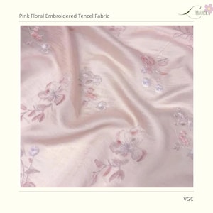 Op de afbeelding: Roze bloemen geborduurde Tencel stof. De stof heeft een zacht, zijdeachtig uiterlijk met een subtiele glans. Delicate bloemenborduursels in roze en grijstinten sieren de stof en cre&euml;ren een romantisch en elegant ontwerp. De stof is gelabeld "AMORRA".
