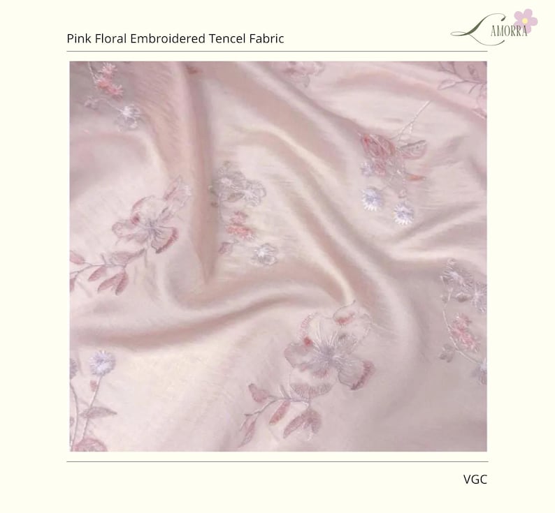 Op de afbeelding: Roze bloemen geborduurde Tencel stof. De stof heeft een zacht, zijdeachtig uiterlijk met een subtiele glans. Delicate bloemenborduursels in roze en grijstinten sieren de stof en cre&euml;ren een romantisch en elegant ontwerp. De stof is gelabeld "AMORRA".