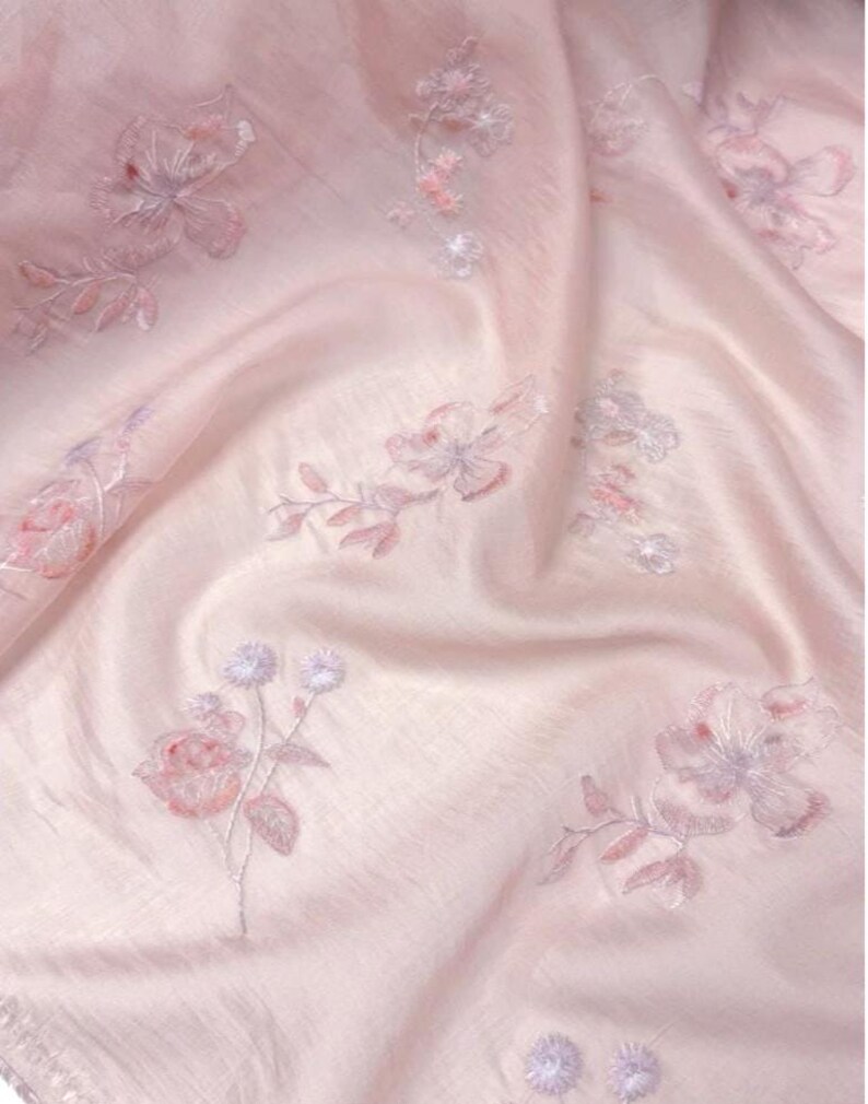 Op de afbeelding: Een zachtroze stof met geborduurde bloemmotieven. De borduursels tonen bloemen en bladeren in roze en lavendel tinten. De stof heeft een zachte, vloeiende uitstraling, met subtiele plooien en highlights, wat een romantische sfeer cre&euml;ert.