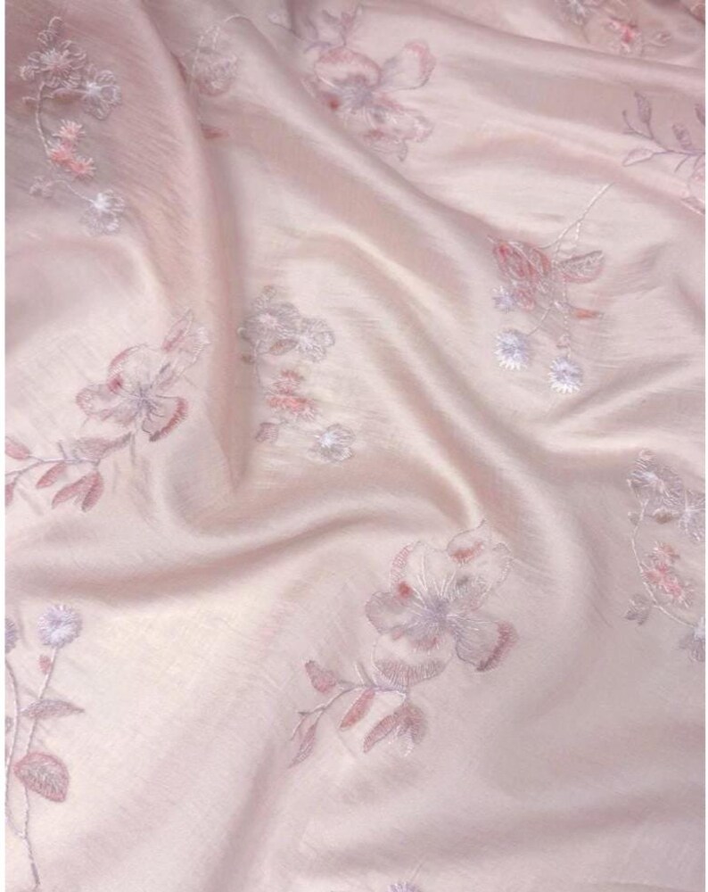 Op de afbeelding: Zachtroze stof met geborduurde bloempatronen. De delicate borduursels tonen bloemen en bladeren in roze en lavendel tinten. De stof heeft een zachte, vloeiende textuur, met subtiele plooien en highlights.