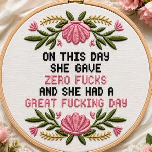 Puede incluir: Un bastidor de bordado circular de madera con tela blanca y el texto bordado: ON THIS DAY SHE GAVE ZERO FUCKS AND SHE HAD A GREAT FUCKING DAY. El diseño presenta motivos florales de color rosa y hojas verdes en la parte superior e inferior.