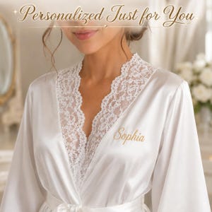 Peut inclure: Un peignoir en satin blanc avec une bordure en dentelle blanche délicate le long de l'encolure en V et des manches longues. Le prénom Sophia est brodé en fil doré sur la poitrine gauche. Le texte Personalized Just for You apparaît en haut.