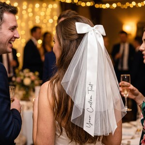 Op de afbeelding: Een witte bruidssluier met een satijnen strik en een lang lint. Op het lint staat de tekst "Your Custom Text". De sluier is gemaakt van een dunne, lichte stof.