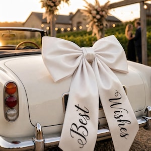 May include: A large white satin bow attached to the rear of a white vintage convertible car. The two long tails of the bow hang down over the number plate area, featuring the black cursive text Best Wishes printed clearly on the fabric surface.