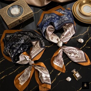 May include: Two square scarves featuring a paisley pattern in black, navy blue, white, and mustard yellow. Each scarf is tied in a knot, displaying a border design with geometric shapes and intricate swirls. The fabric appears smooth and lightweight.