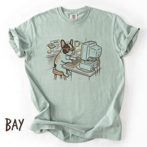 May include: A sage green short-sleeve t-shirt featuring a graphic illustration of a French bulldog wearing glasses, sitting on a stool at a desk with a vintage computer, keyboard, and a document labeled DOGE, with a coffee mug nearby.