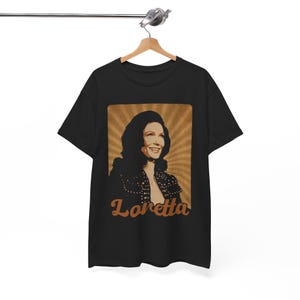 May include: A black short-sleeve t-shirt featuring a rectangular graphic print of a smiling person with dark hair. The background of the print has brown sunburst rays, and the name Loretta is written in a brown cursive font below the portrait.