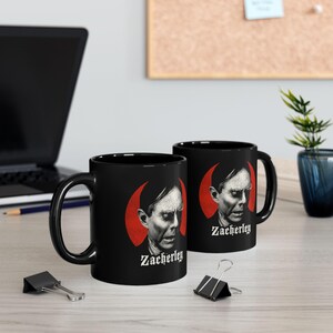 Puede incluir: Dos tazas de cerámica negra con un retrato monocromático de Zacherley centrado sobre un fondo circular rojo intenso. El nombre Zacherley está impreso en una fuente gótica blanca justo debajo del retrato en cada taza.