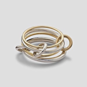 Puede incluir: Un anillo de múltiples bandas que presenta una serie de aros circulares entrelazados en tonos alternos de oro y plata pulidos. El diseño consta de varios anillos de metal finos y lisos que están conectados y superpuestos en un grupo.