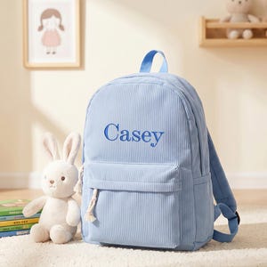 Könnte beinhalten: Ein hellblauer Rucksack aus Cord mit vertikaler Rippenstruktur und dem Namen Casey, der in dunkelblauem Garn auf die Vordertasche gestickt ist. Der Rucksack verfügt über einen Tragegriff, verstellbare Schultergurte und einen weißen geknoteten Reißverschluss.