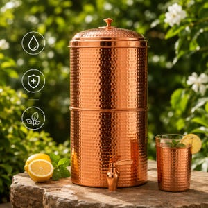 May include: A copper water filter with a hammered texture, a matching glass, and a spigot. The filter is cylindrical with a lid and sits on a stone surface. A glass of water with a lemon slice and mint is next to it.